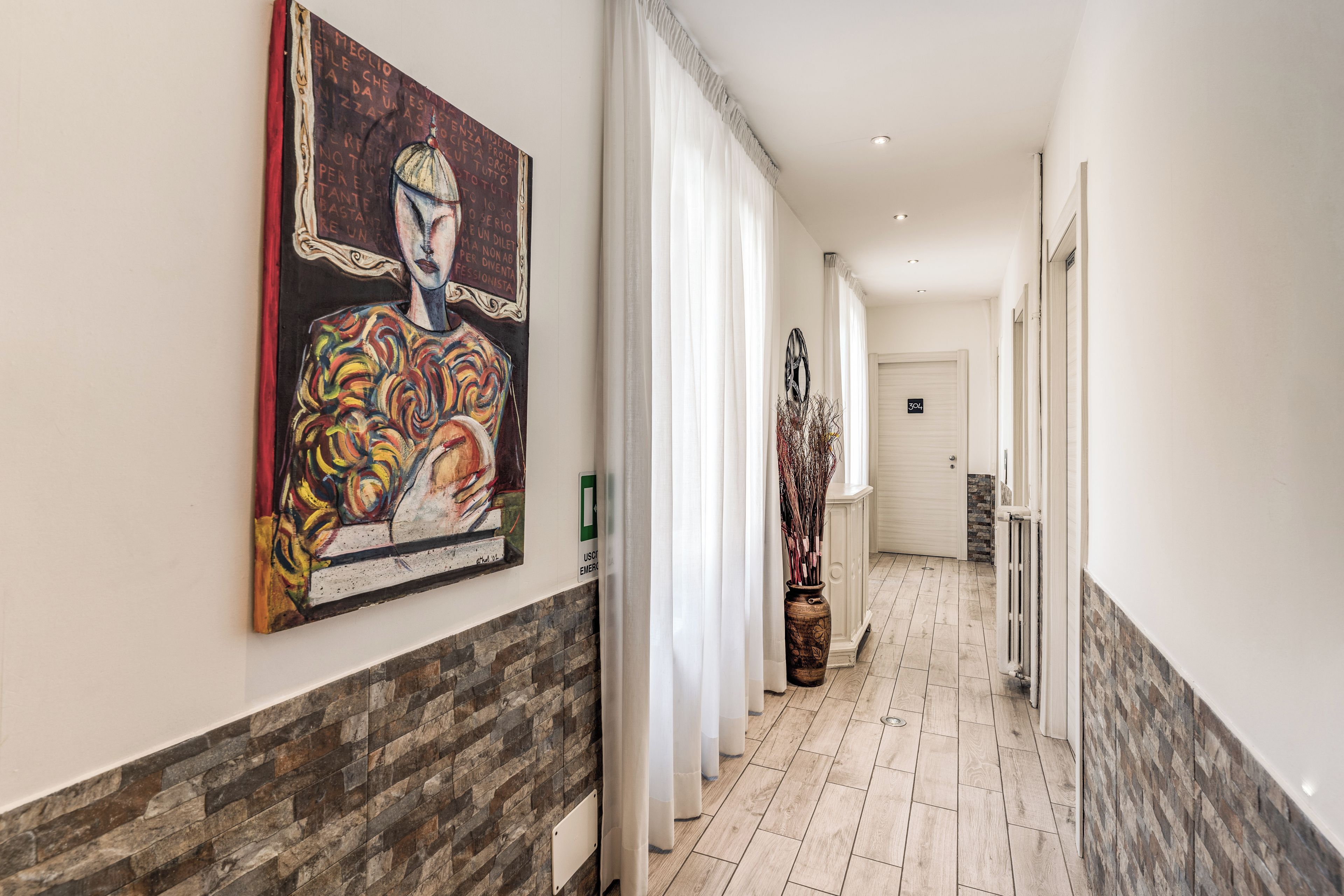 Een hotelgang met een groot abstract schilderij aan de muur, een witte deur aan het einde, en een wand met een stenen patroon.