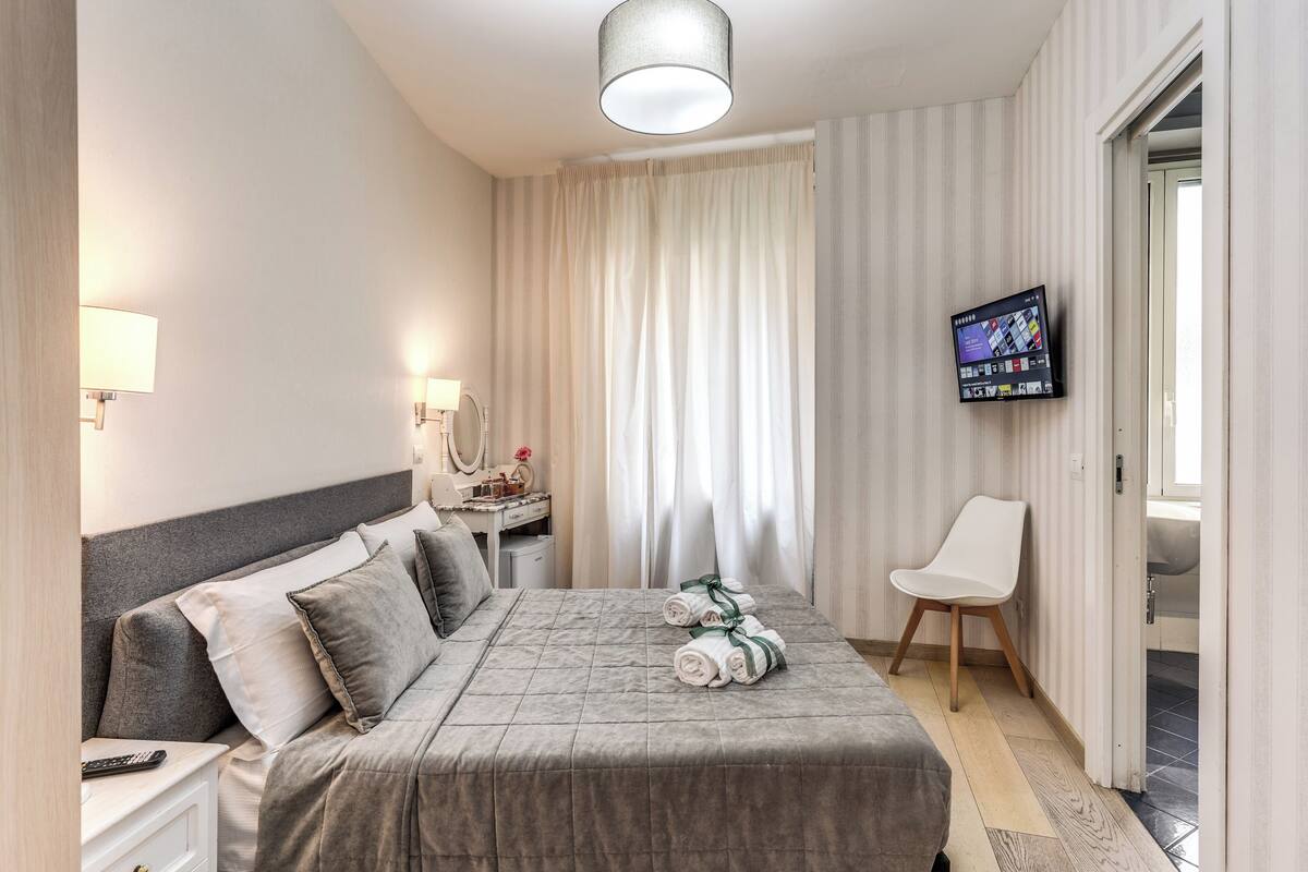 A modern hotel room with a neatly made bed, a television mounted on the wall, and a chair by the window.