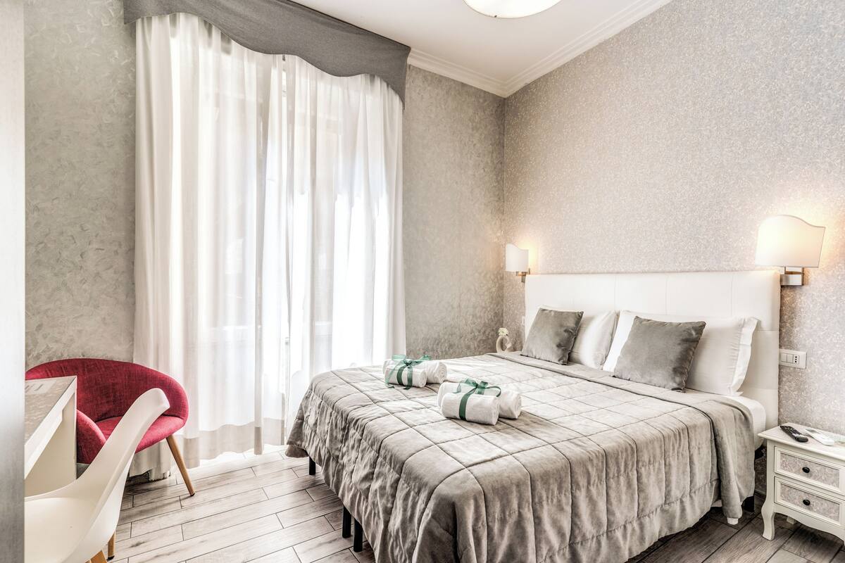 A modern hotel room with a large bed, a red armchair, a white chair, and a nightstand with a lamp.
