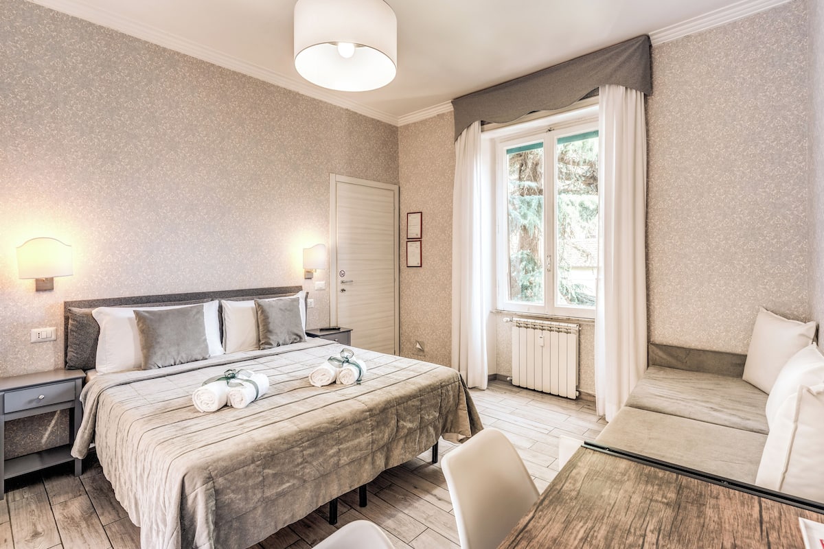 A hotel room with a bed, bedside tables, a desk, a chair, a window with curtains, and a radiator.