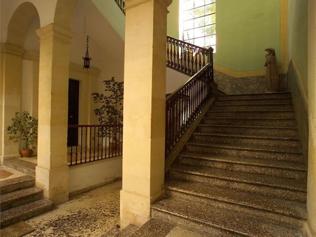 Un escalier avec des marches en pierre et une rampe en fer forgé, menant à un palier où se trouvent une porte et un pot de fleurs.