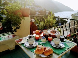 Un balcón con una mesa dispuesta para el desayuno, que incluye cruasanes, café y jugo, con vistas a un pueblo costero.