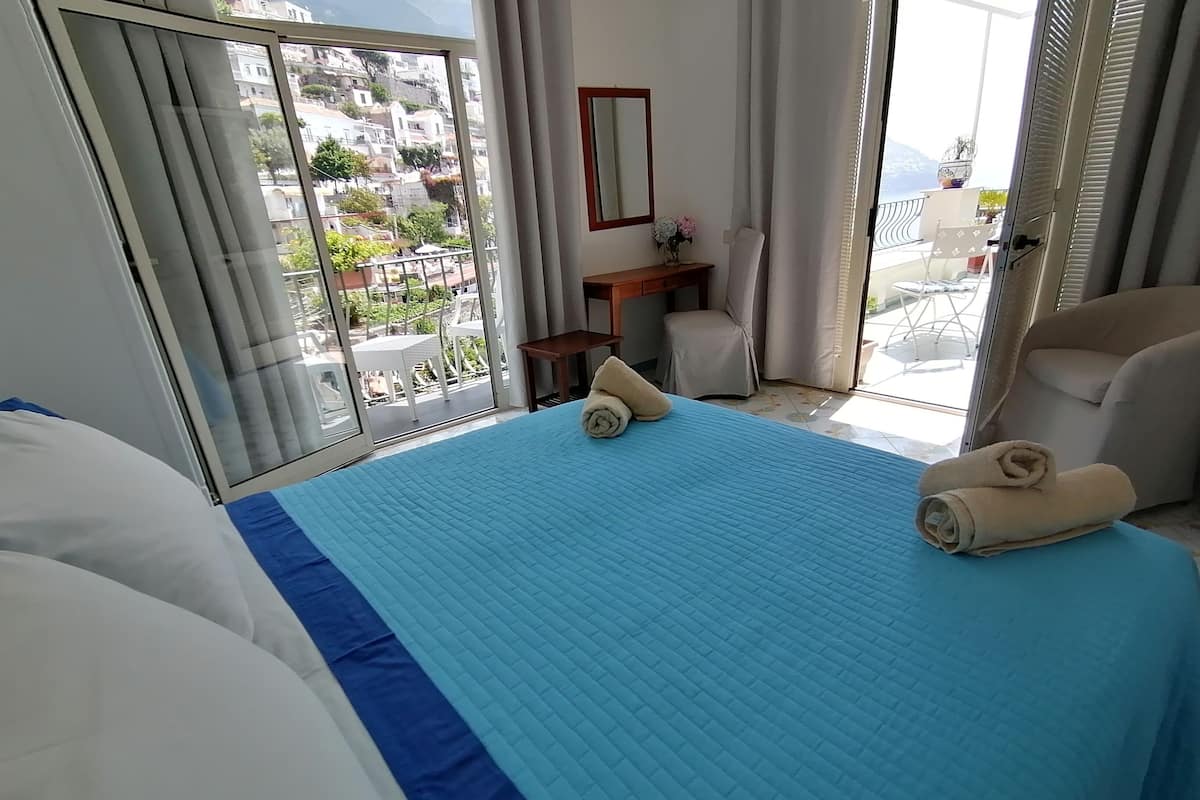 Quarto de hotel com cama, poltrona, mesinha, espelho e vista da cidade através de grandes janelas.