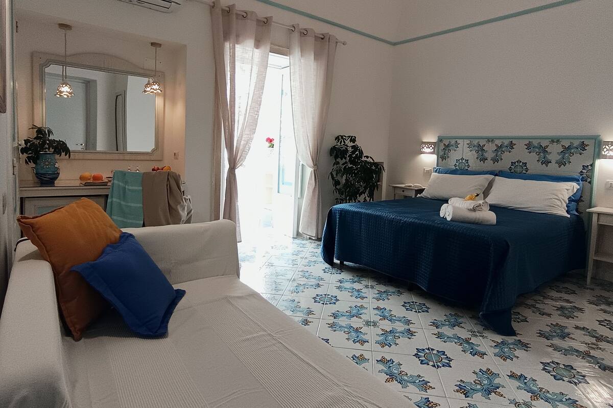 Quarto de hotel com cama, sofá, espelho, mesa de cabeceira e planta em vaso.
