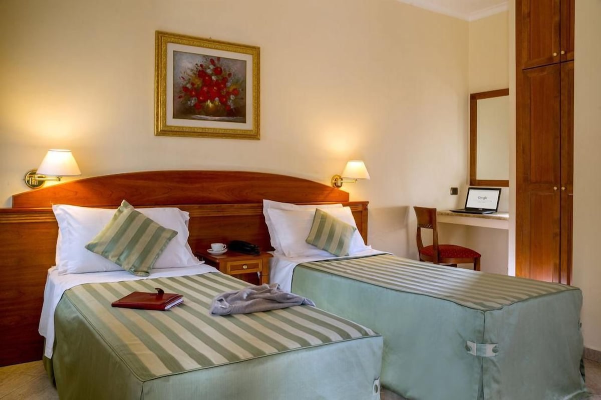A hotel room with two beds, a wooden headboard, bedside lamps, a desk with a laptop, a chair, and a painting on the wall.
