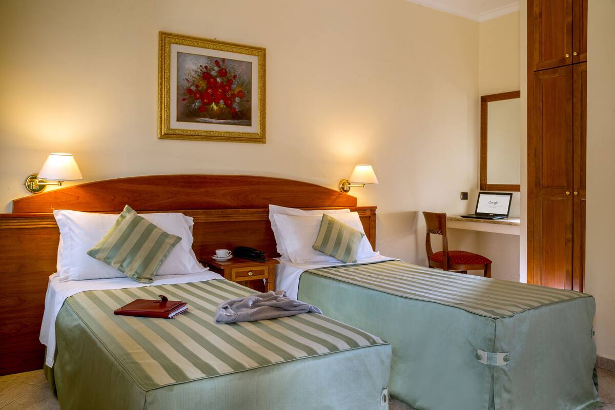 A hotel room with two single beds, a wooden headboard, bedside lamps, a framed painting, a desk with a laptop, and a chair.