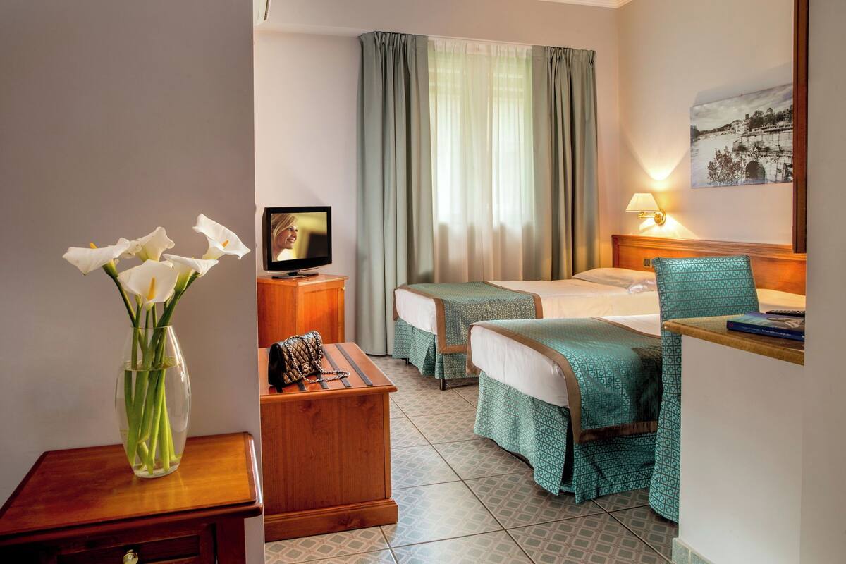 A hotel room with two beds, a TV, a vase with flowers, a bedside table, and a lamp.