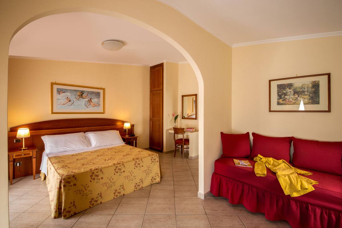 A hotel room with a bed, a red sofa, a desk, and two paintings on the walls.