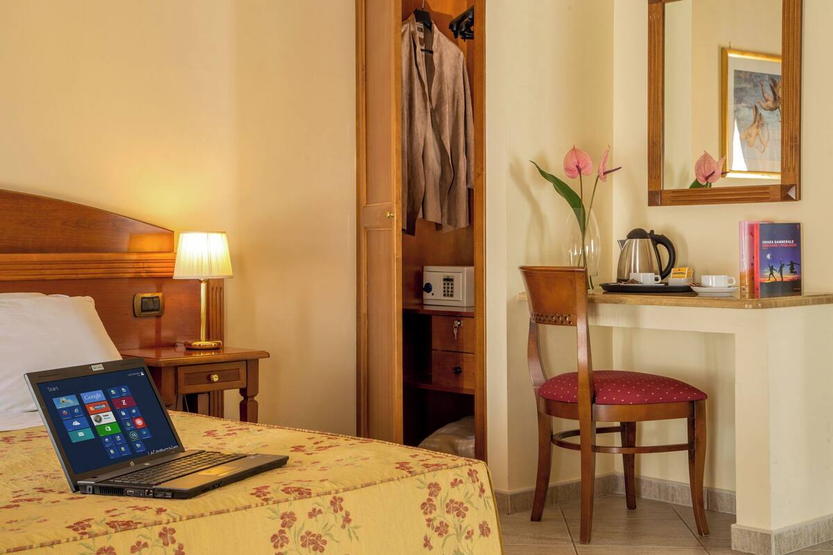 A hotel room with a bed, a laptop on the bed, a desk with a chair, a mirror, a vase with flowers, and a coffee maker.