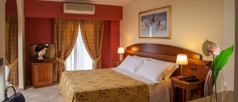 A hotel room with a bed, bedside lamps, a desk with a telephone, a vase with a plant, and a painting on the wall.