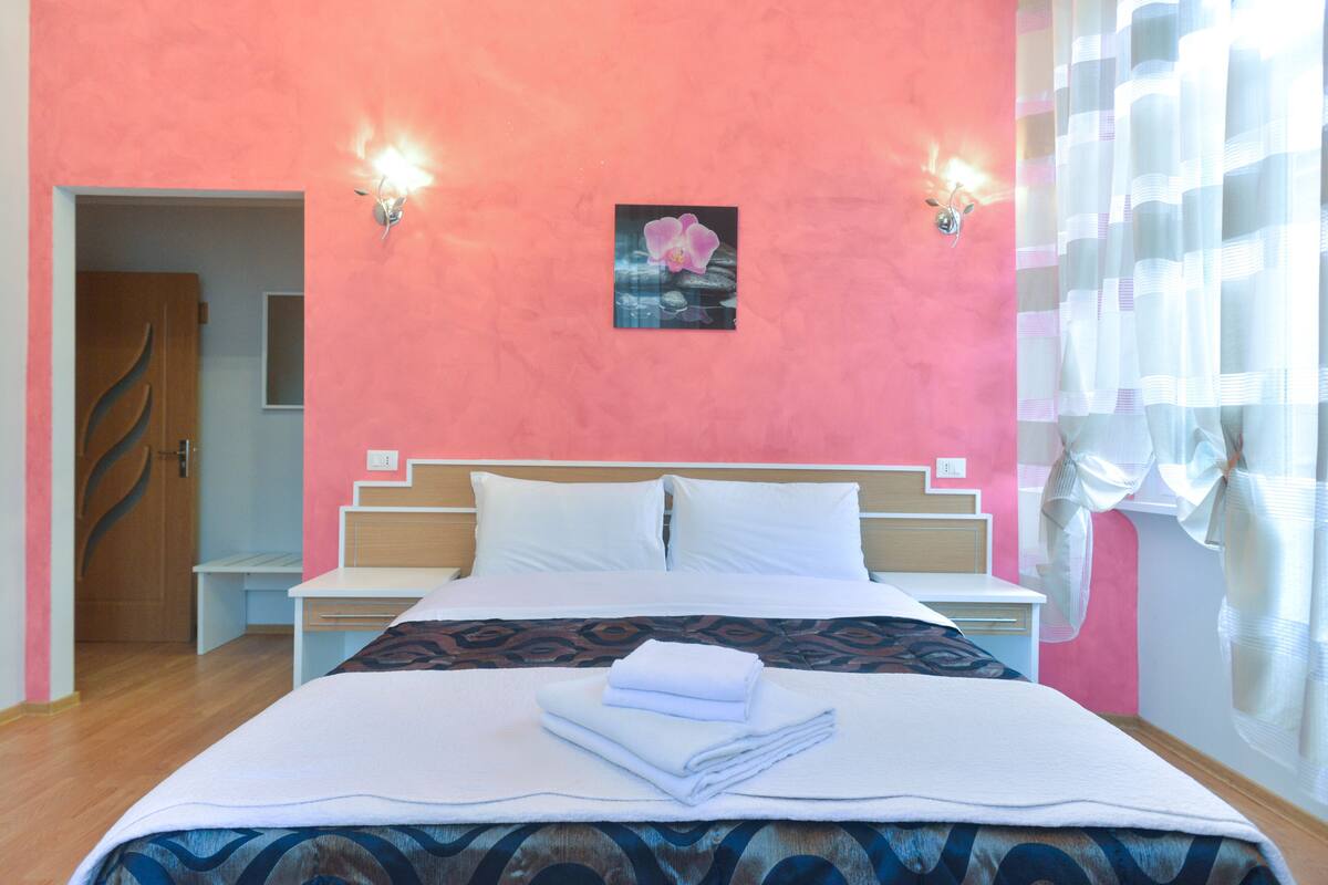 A hotel room with a bed, bedside tables, a nightstand, a painting, and a window with curtains.