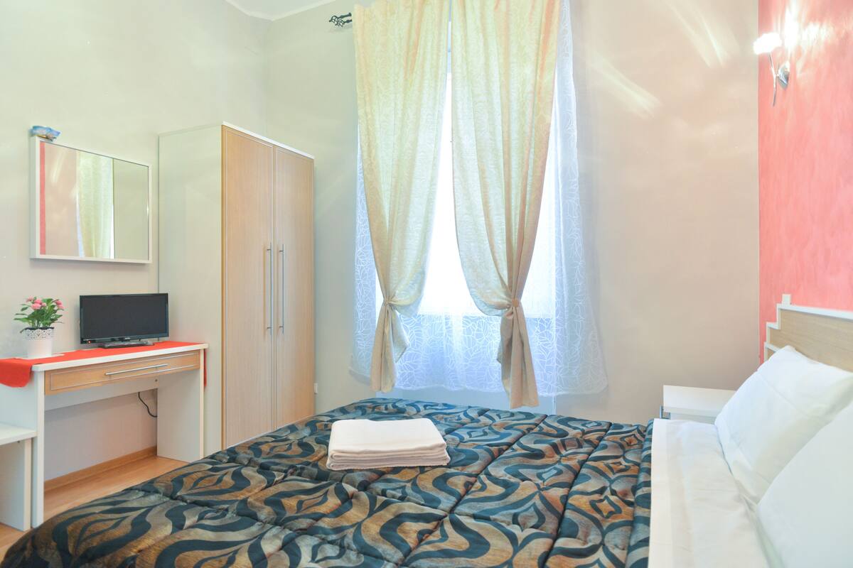 A bedroom with a bed, a desk with a television, a wardrobe, and a window with curtains.