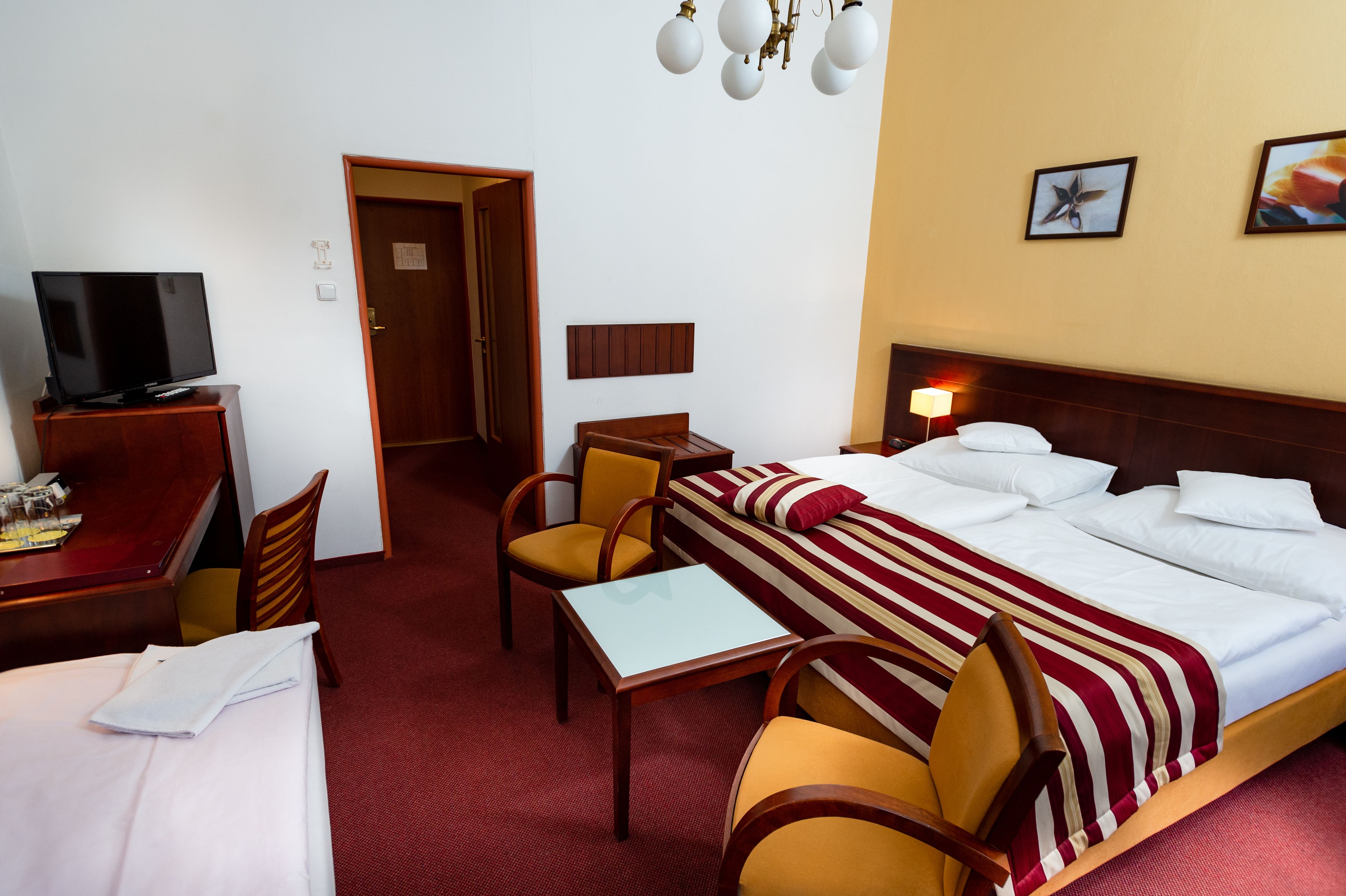 A hotel room with a bed, a desk with a TV, a chair, a table, and a door leading to another room.