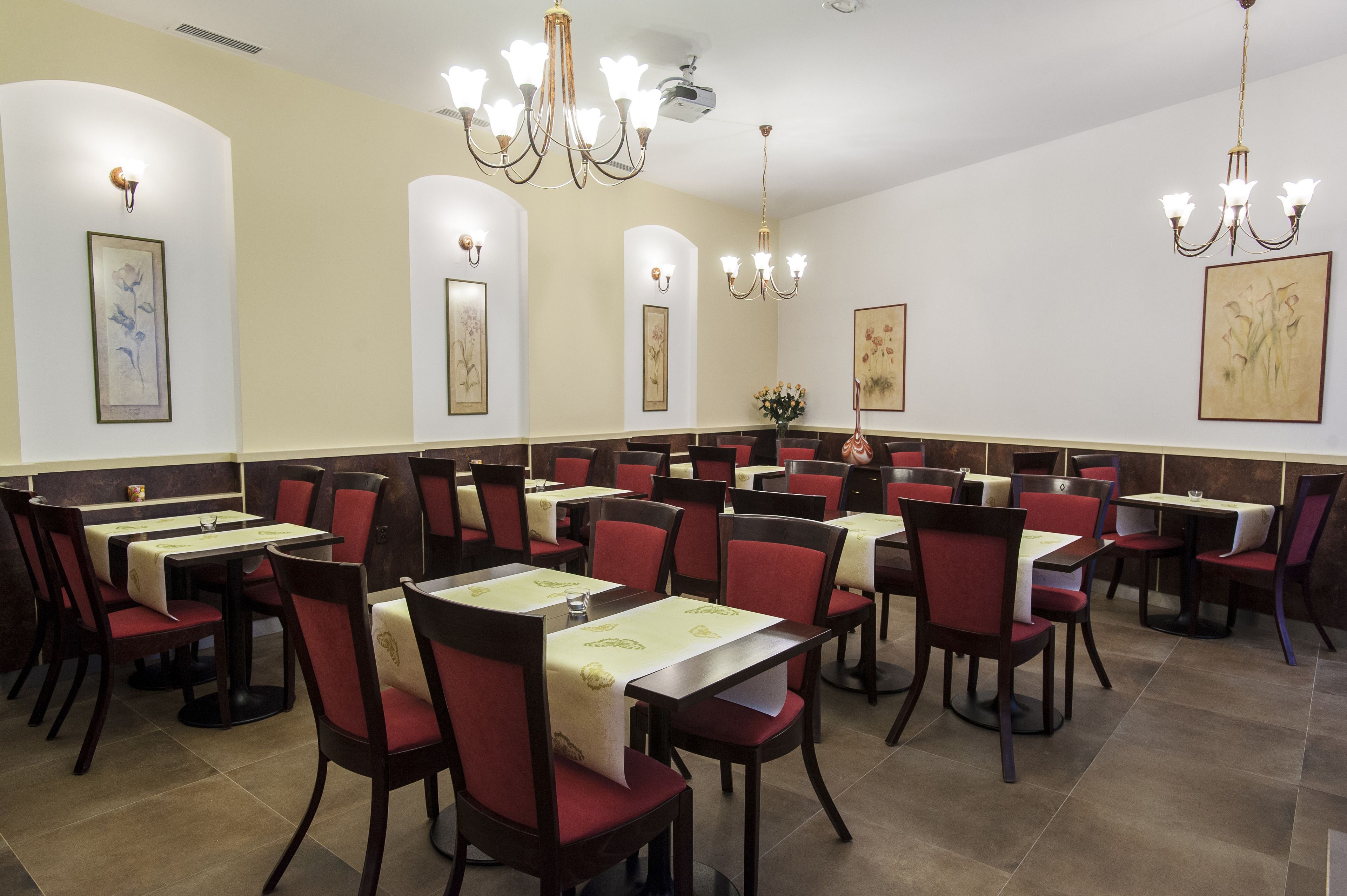 A restaurant with multiple tables and chairs, each with a tablecloth. There are wall-mounted light fixtures and framed artwork on the walls.