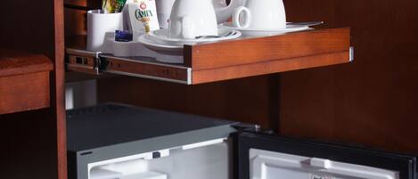 A compact refrigerator with a shelf above containing a coffee maker, cups, and a small container. Inside, there are bottles of beer and cans of soda.