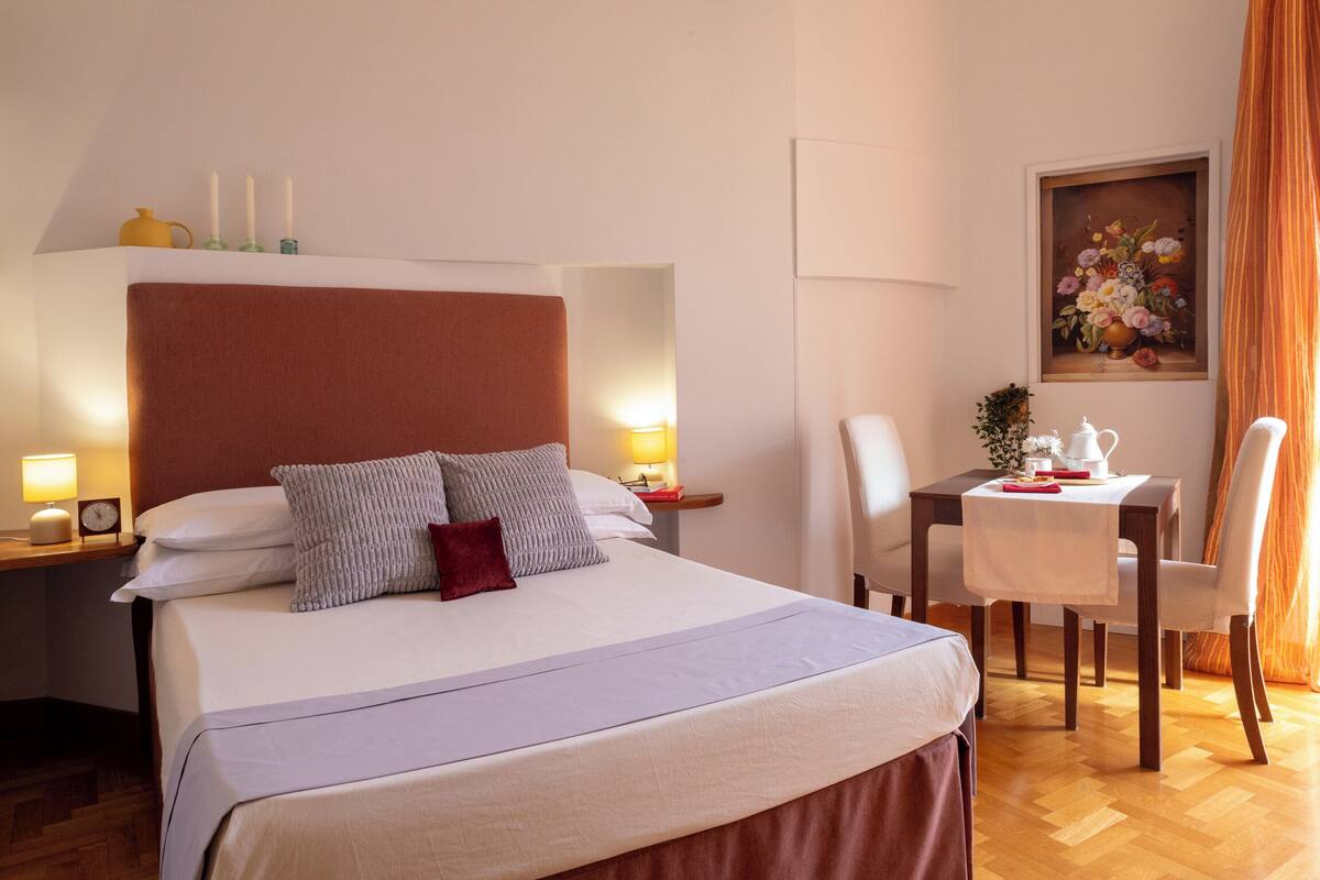 A hotel room with a large bed, bedside tables with lamps, a small table with a teapot, and a painting on the wall.