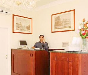 A reception desk with framed pictures on the wall, a computer, and a vase with flowers. - Hotel Chicago (Rome)
