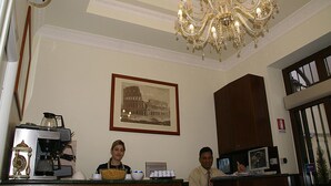 A reception area with a chandelier, a framed picture of the Colosseum, a coffee machine, and a counter with a person standing behind it.