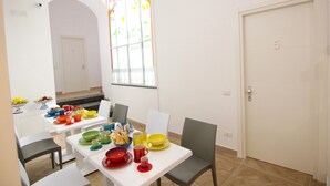 Un coin repas avec une table blanche dressée pour quatre personnes, entourée de chaises. Il y a une fenêtre donnant sur des arbres et une porte portant le numéro 5.