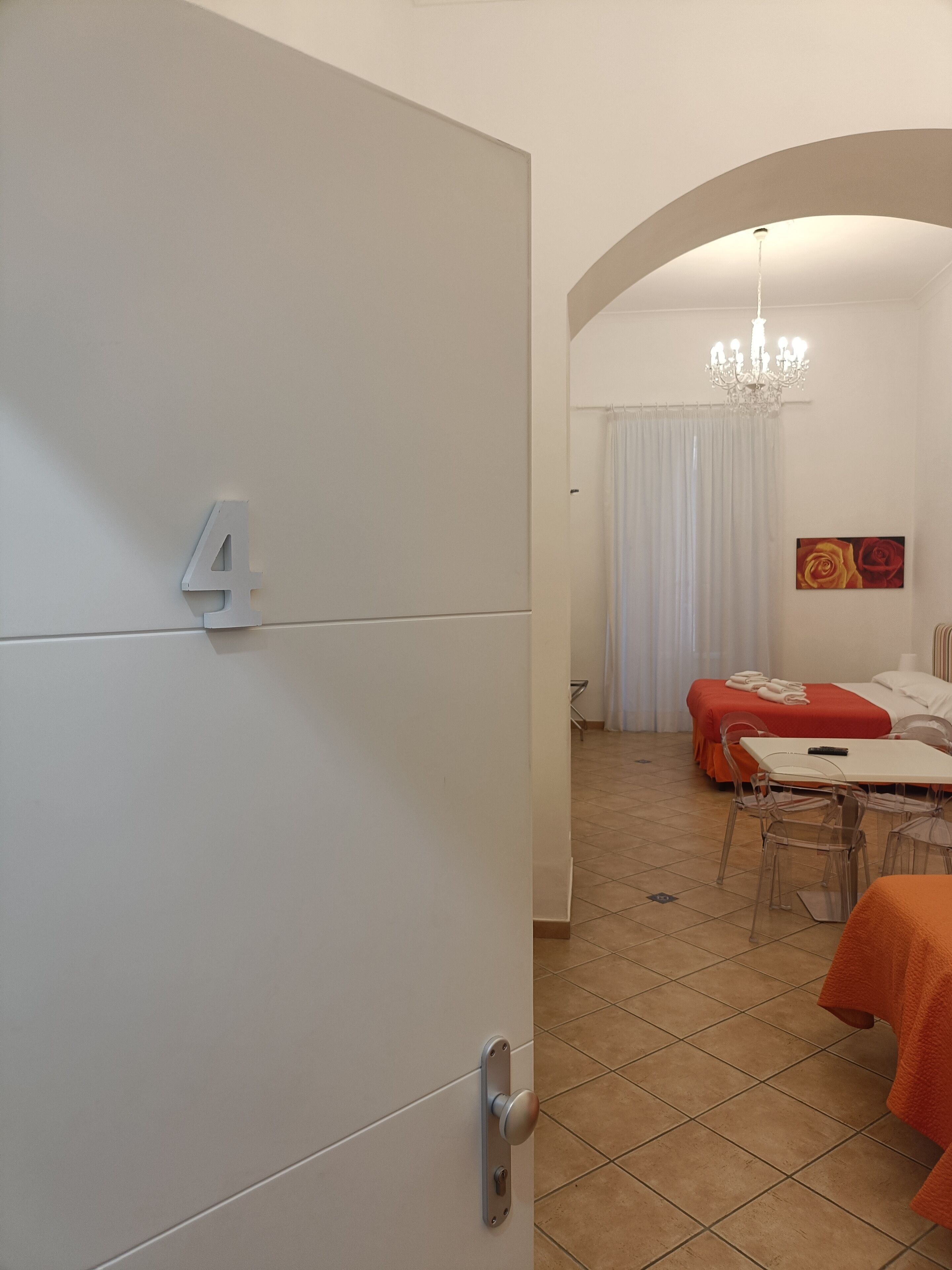 Habitación de hotel con el número 4 en la puerta, una cama con una manta roja, una mesa de comedor con sillas y una araña.