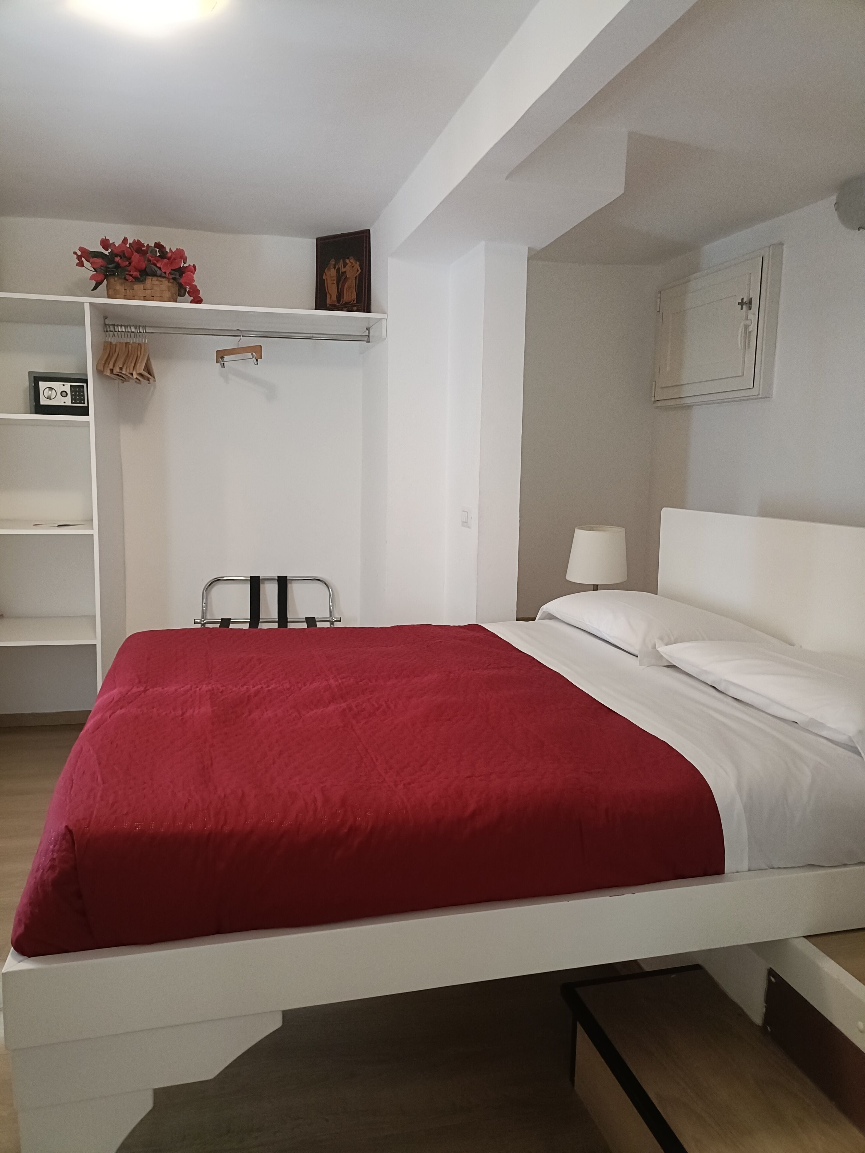 A bedroom with a white bed, red bedding, a white wardrobe, and a small table with a lamp.