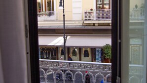 Une vue depuis un balcon doté d’une rambarde en métal, une rue avec des piétons, et des immeubles avec des balcons et des auvents.