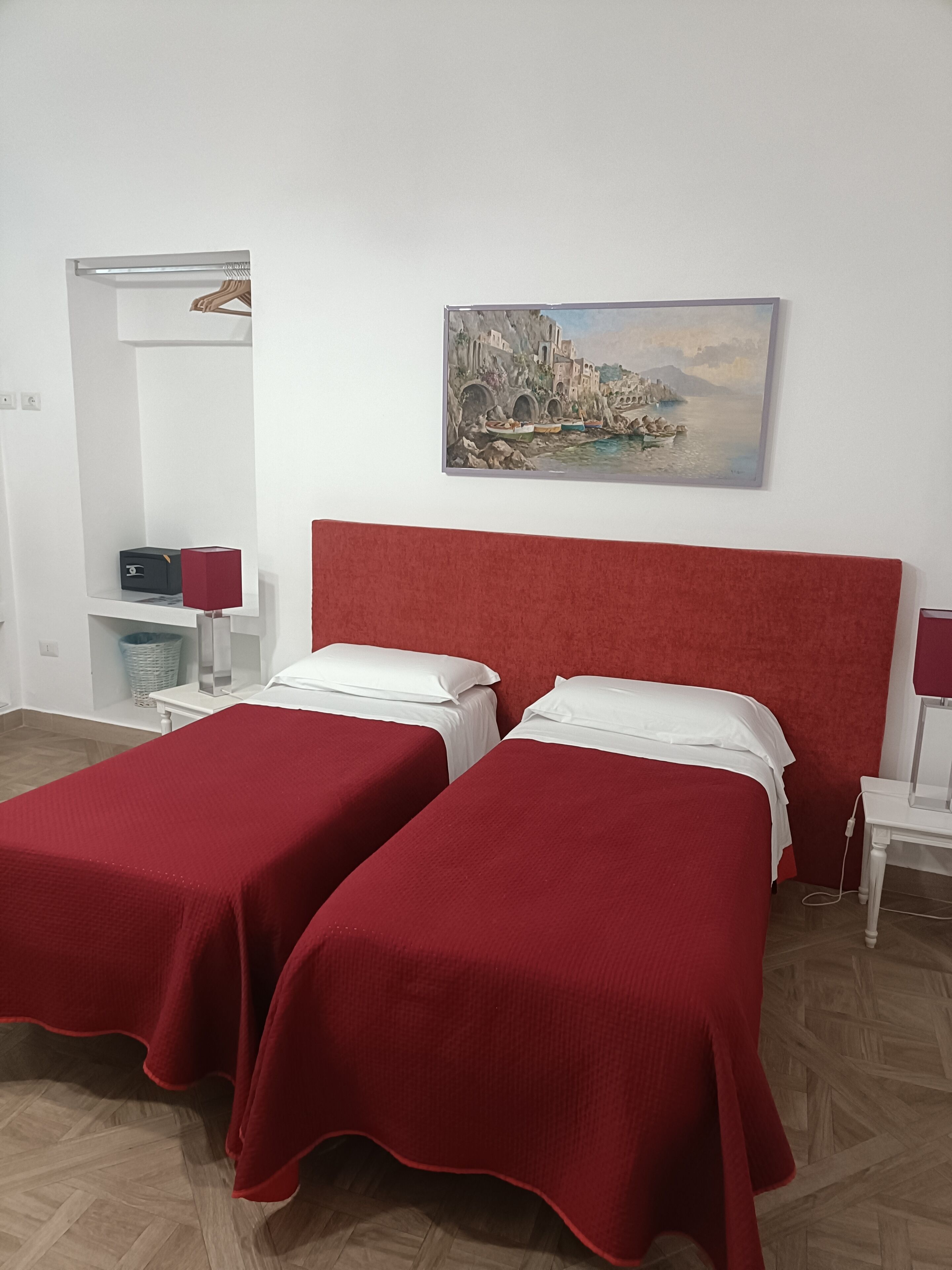 Une chambre d’hôtel avec deux lits simples, une tête de lit rouge, un tableau représentant un paysage côtier, une table de chevet blanche et une lampe rouge.