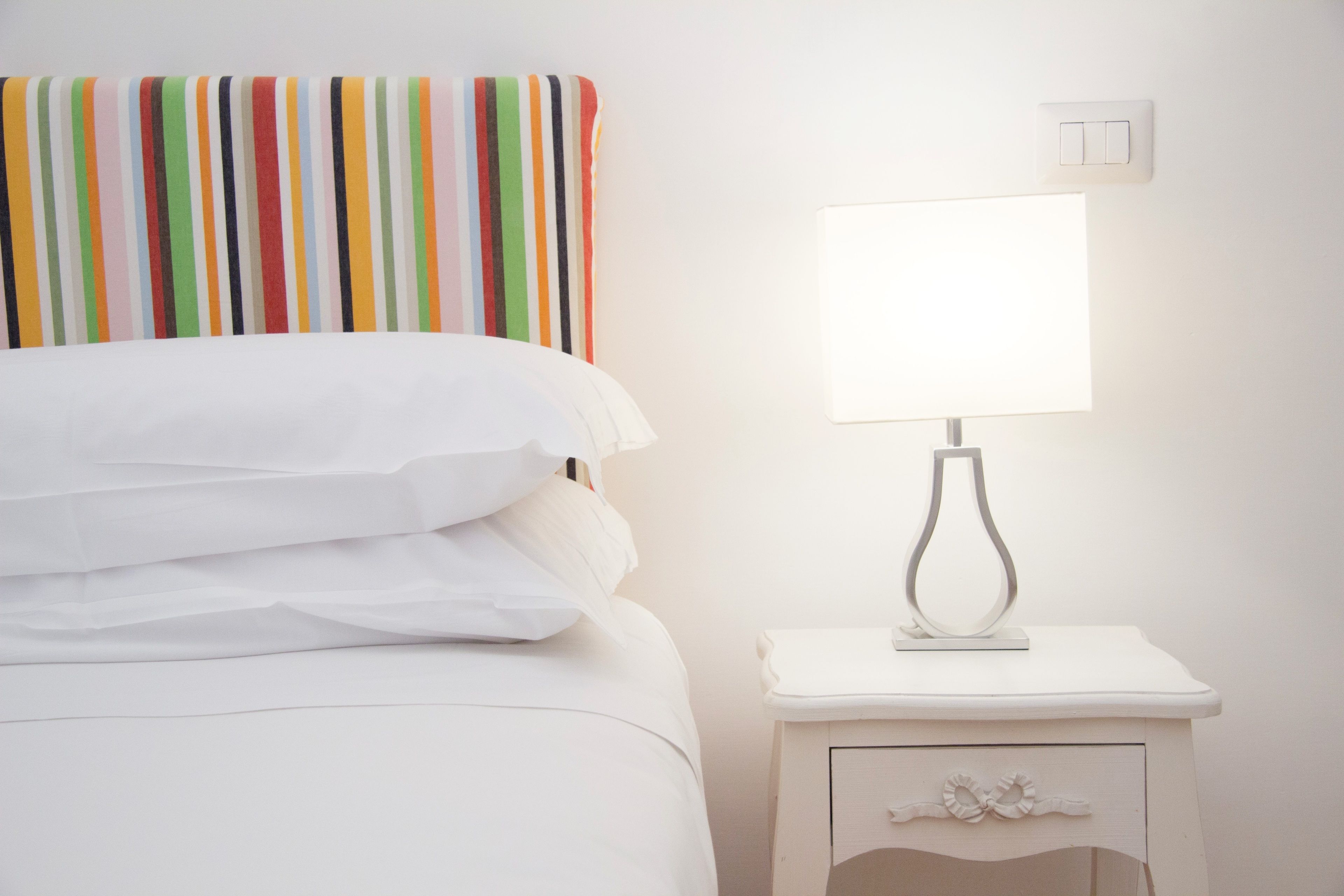 A neatly made bed with white linens, a bedside table with a lamp, and a colorful striped headboard.