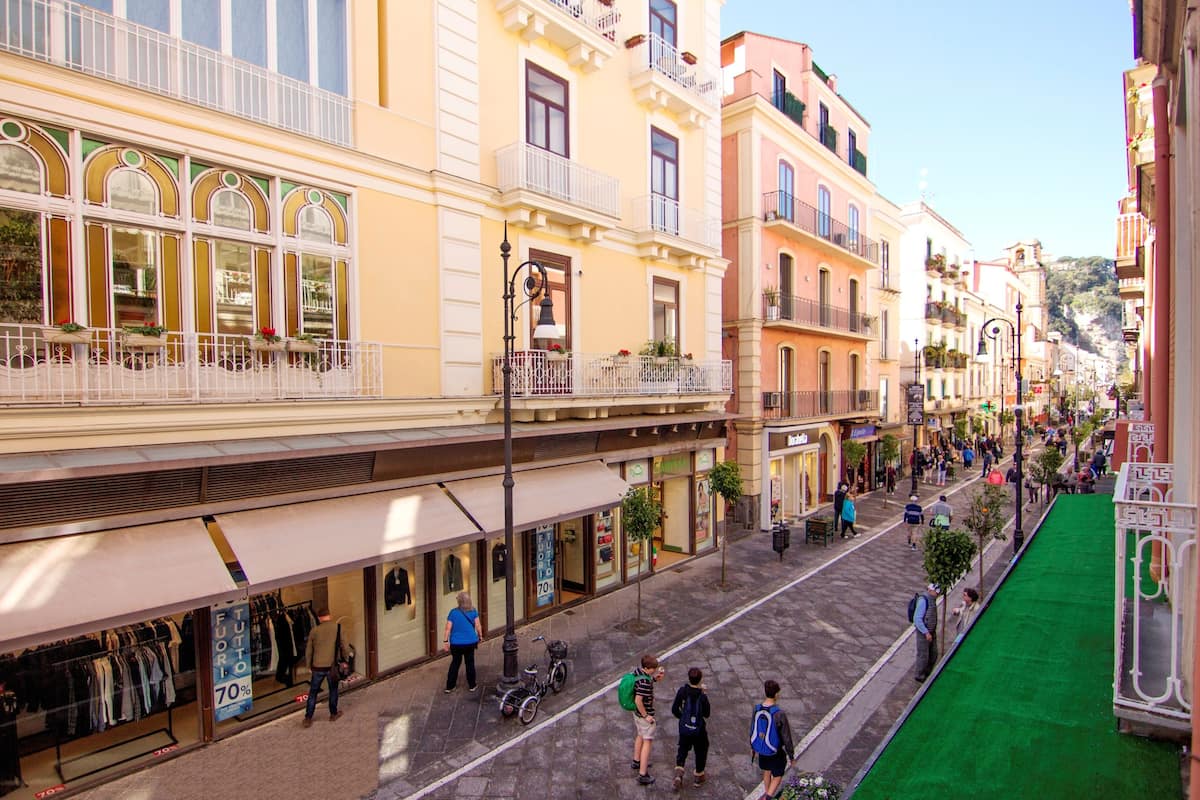Un carrer amb botigues i balcons, una tenda amb teulada verda, i persones caminant.