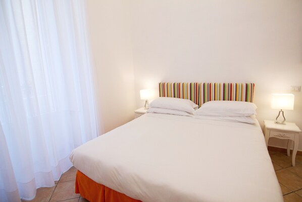 Habitación de hotel con una cama grande, ropa de cama blanca, cabecera a rayas, dos mesitas de noche con lámparas y una ventana con cortinas de gasa.