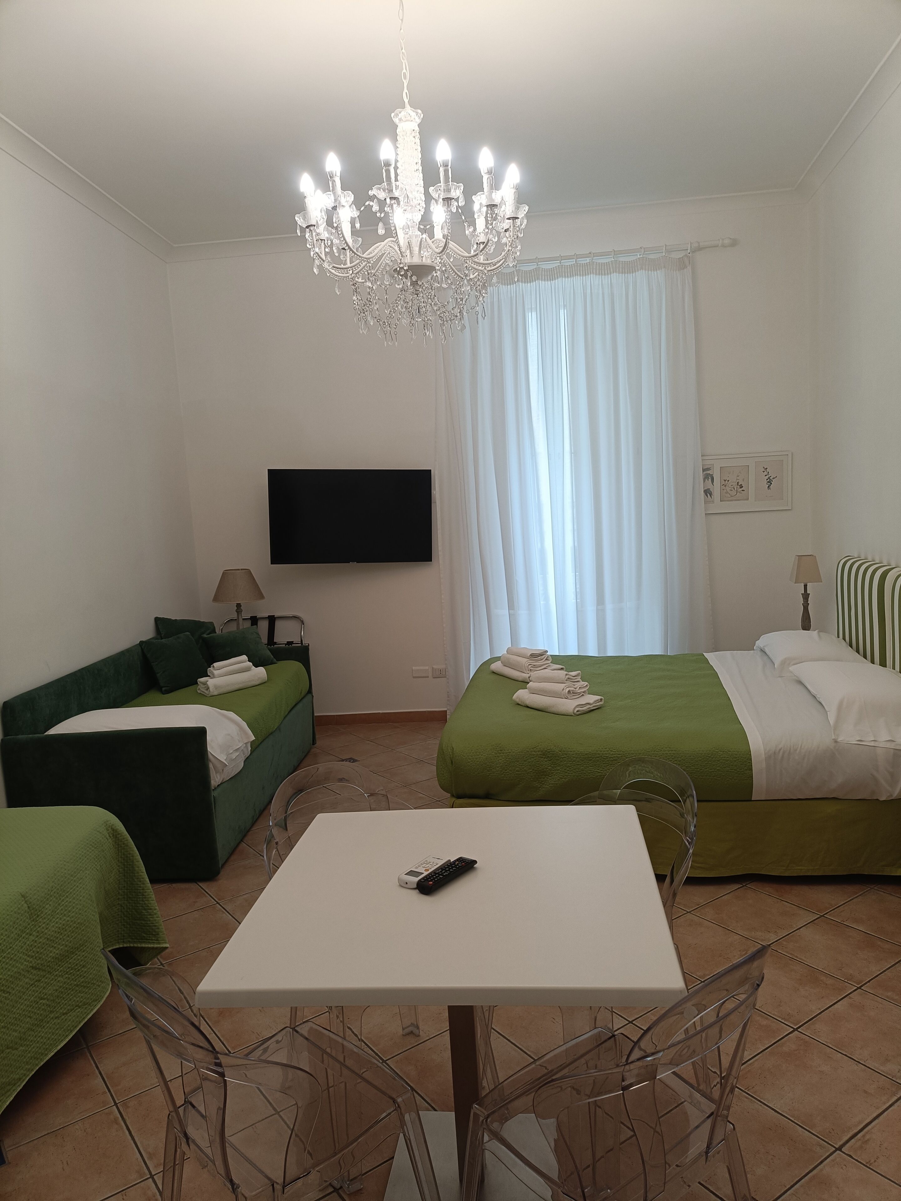 Une chambre d’hôtel avec un canapé vert, un lit avec une literie verte, une table blanche, un lustre, un téléviseur à écran plat et une fenêtre avec des rideaux blancs.