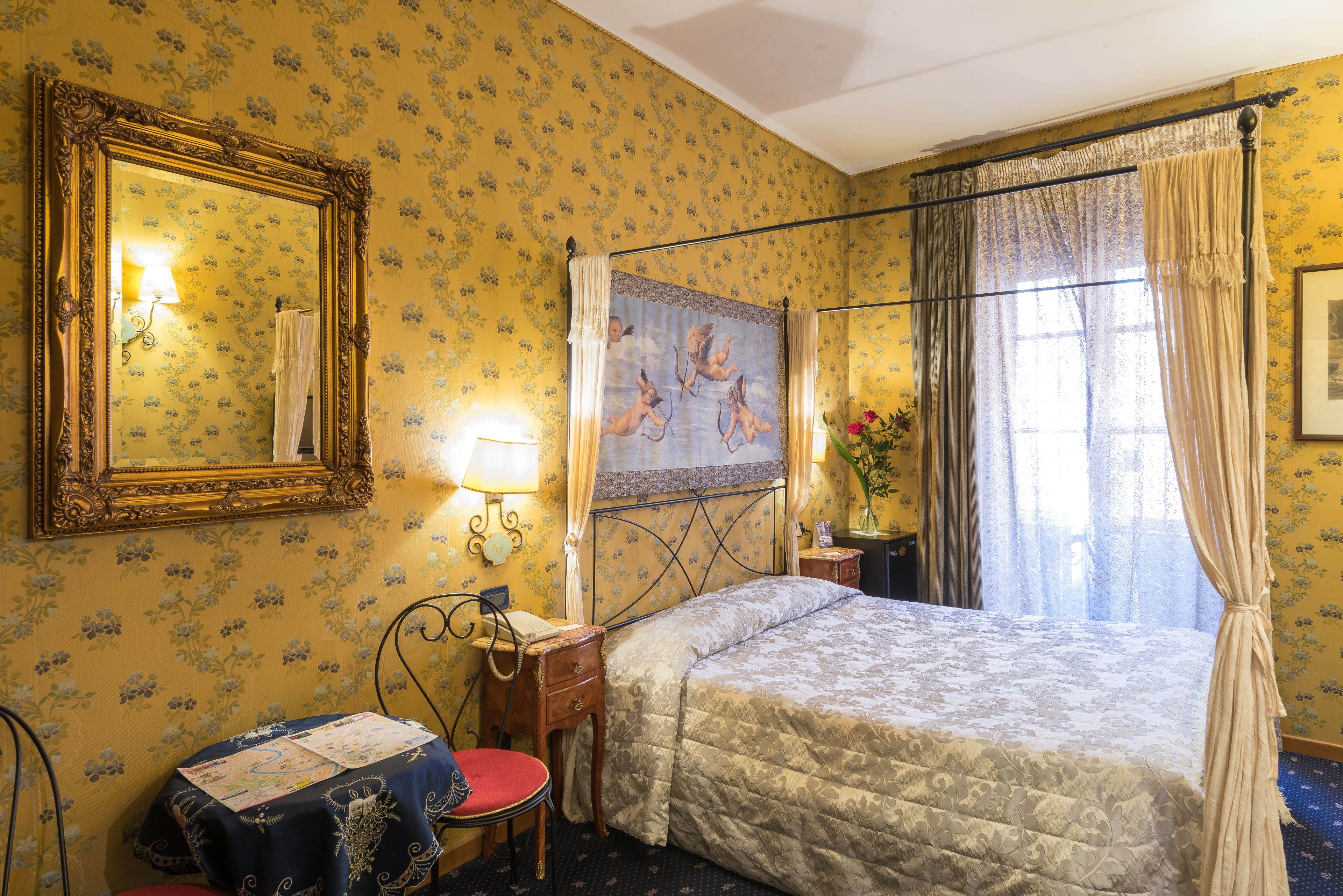 A hotel room with a four-poster bed, a large mirror, floral wallpaper, a small table with a lamp, and a window with curtains.