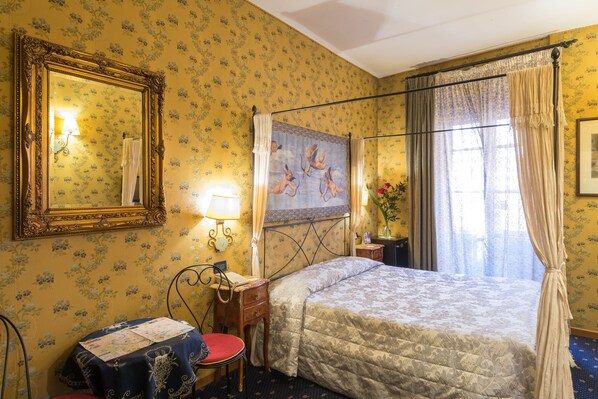 A hotel room with a four-poster bed, a large mirror, floral wallpaper, a small table with a lamp, and a window with curtains. - Residenza Ave Roma (Rome)