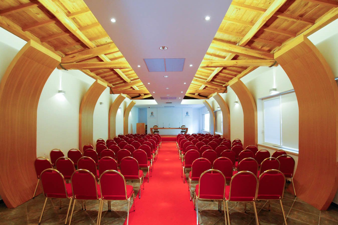 Ein Konferenzraum mit roten Stühlen, die in Reihen angeordnet sind, einem roten Teppich, Holzbalken an der Decke und einem Rednerpult im vorderen Bereich.