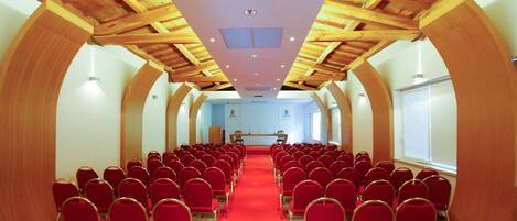 Een congreszaal met rode stoelen in rijen, een rood tapijt, houten balken in het plafond en een spreekgestalte aan de voorzijde.