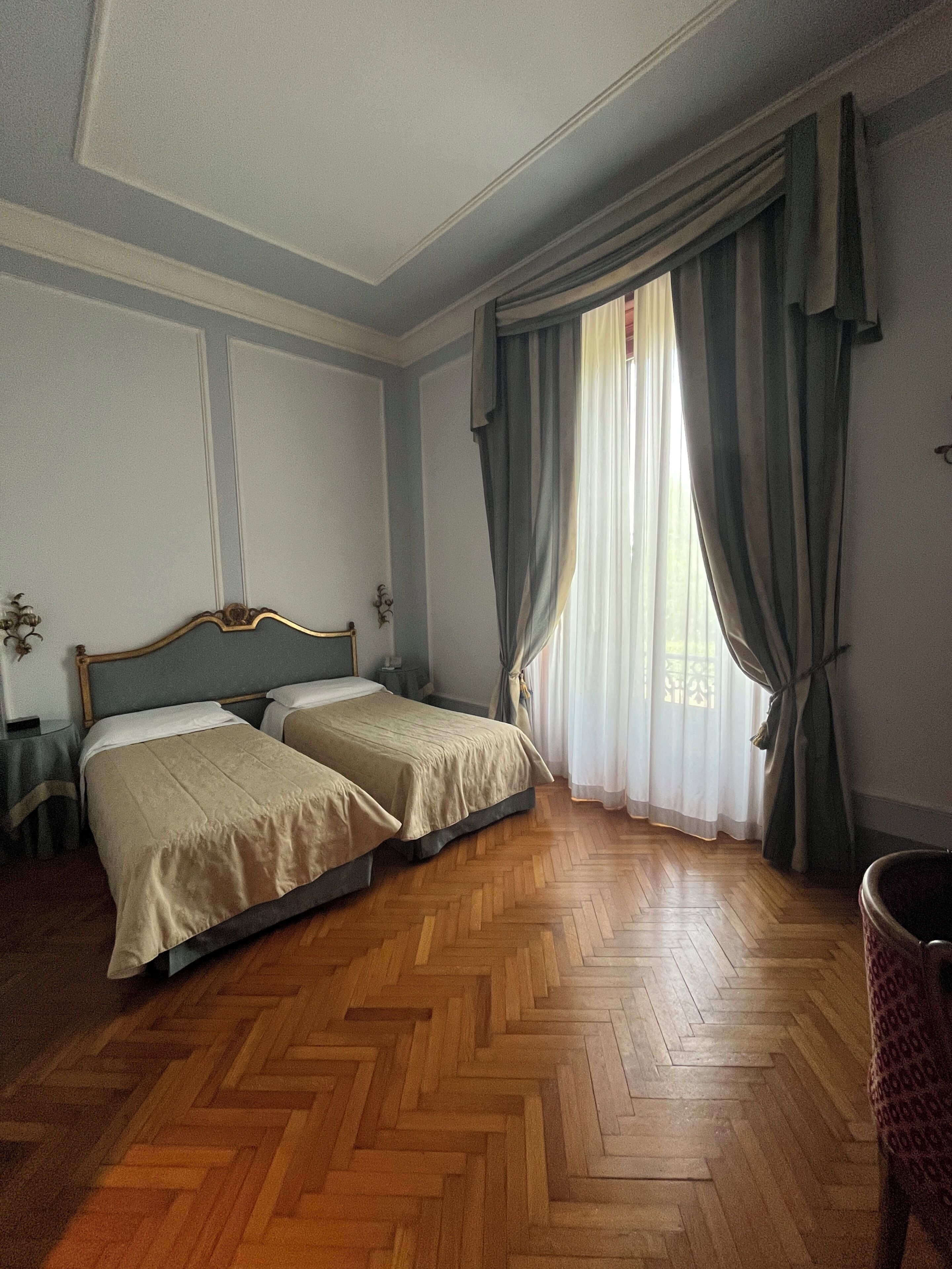 A room with two beds, a wooden herringbone floor, a window with curtains, and a wall-mounted lamp.