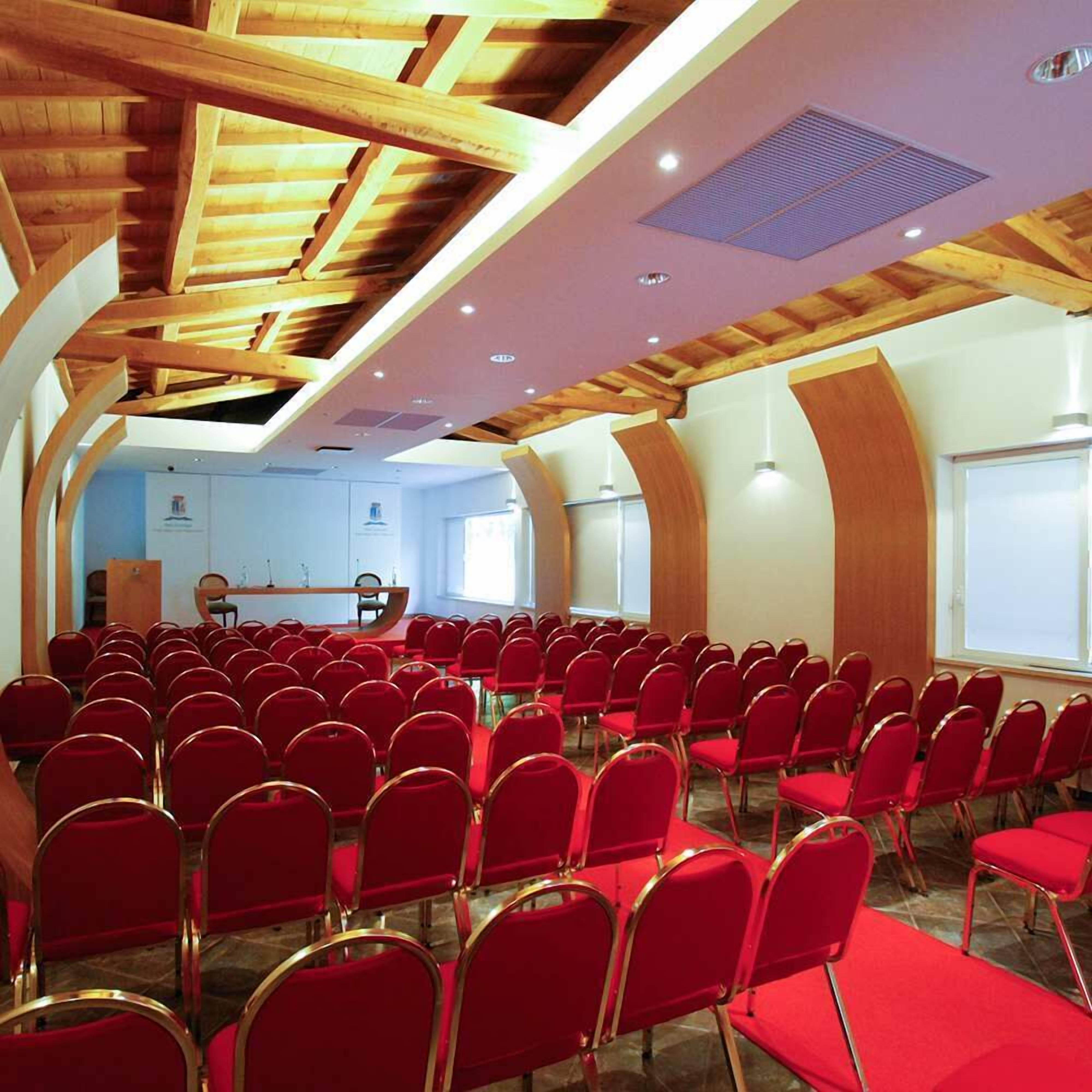 Een congreszaal met rode stoelen in rijen, een podium en een hoog houten plafond met dakramen.