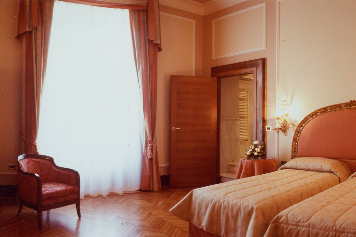 Quarto de hotel com duas camas, uma cadeira de madeira, uma porta e uma mesa com flores.