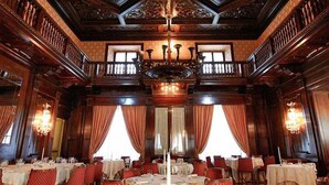 Un amplio salón comedor con techos de madera ornamentada, candelabros y sillas tapizadas en rojo. Las mesas están dispuestas para el servicio.