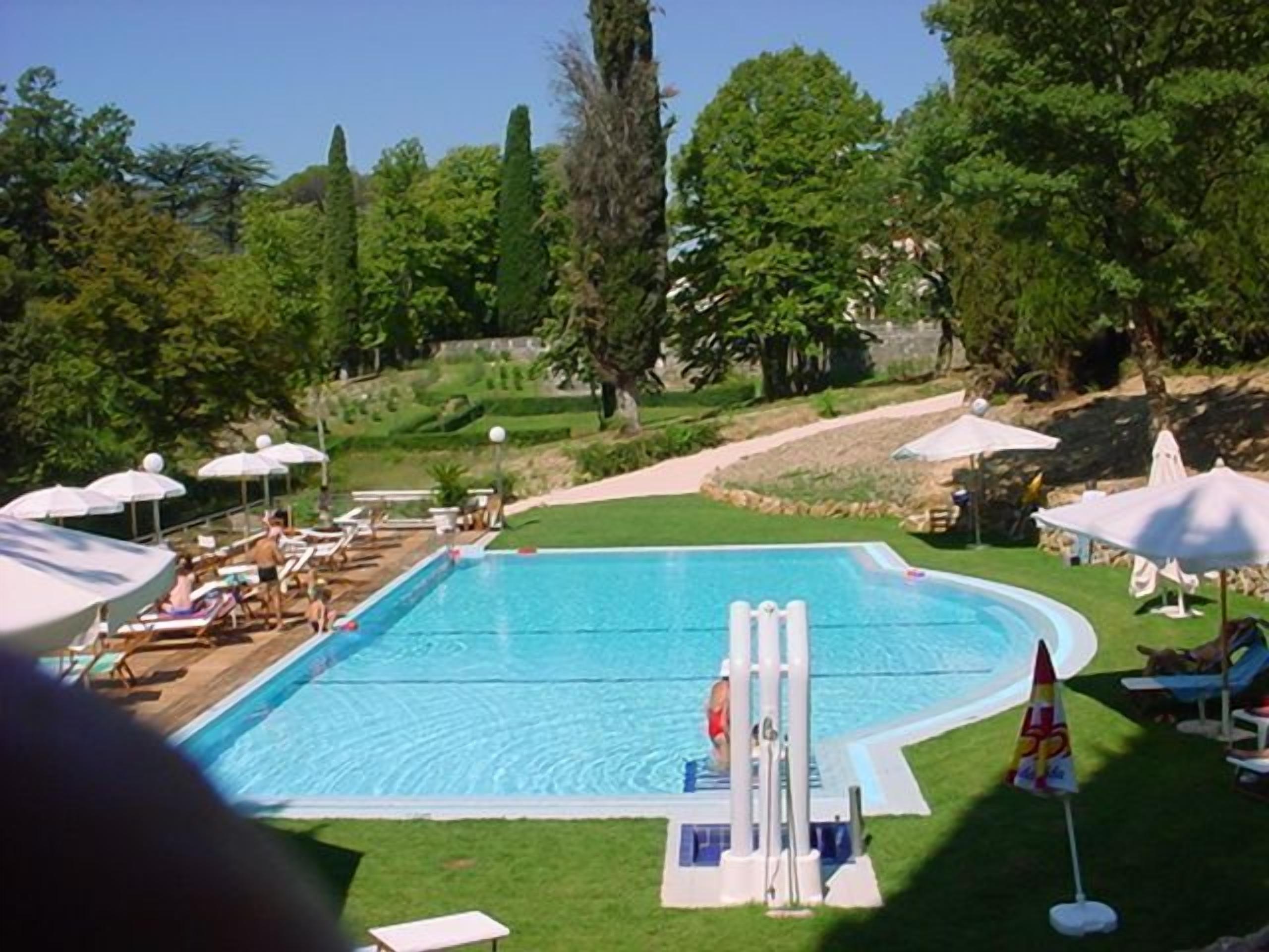 Ein Schwimmbecken mit Liegestühlen und Sonnenschirmen, umgeben von Bäumen und einem gepflegten Rasen.