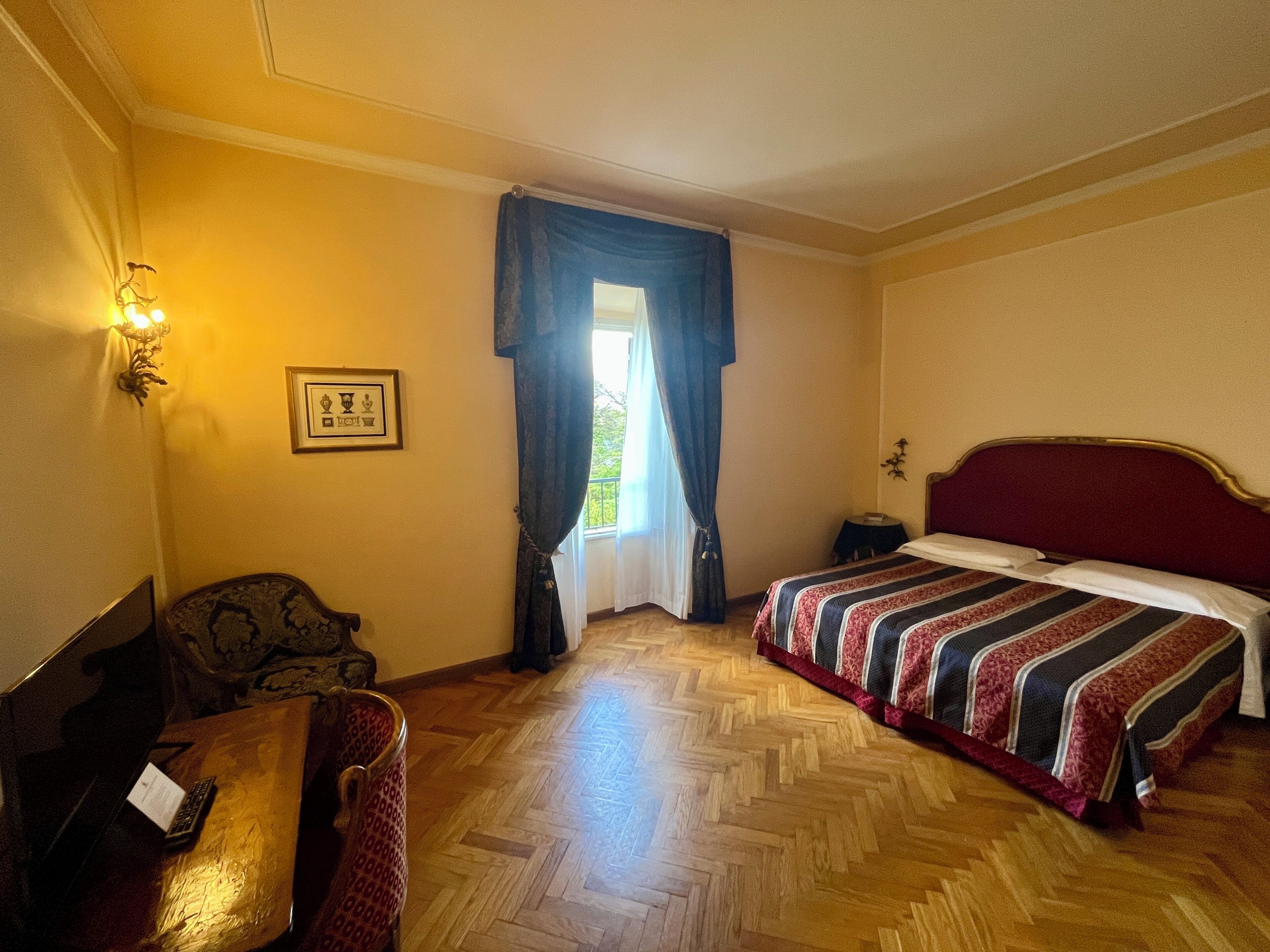 Una habitación de hotel con cama, escritorio, silla, lámpara y ventana con cortinas.