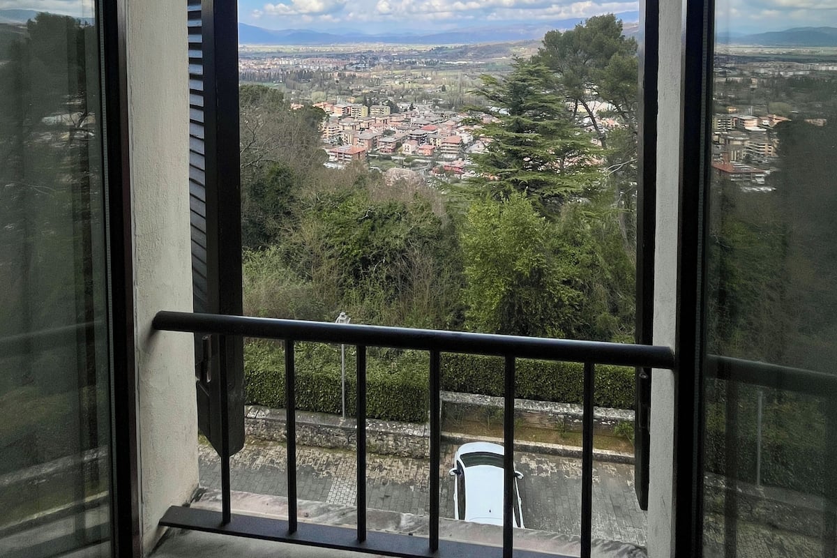 Vista através de uma janela com grade de metal, mostrando um horizonte urbano com edifícios e árvores.