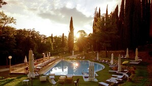 Un área de piscina con sillones de descanso y sombrillas, rodeada de árboles y un cielo despejado.