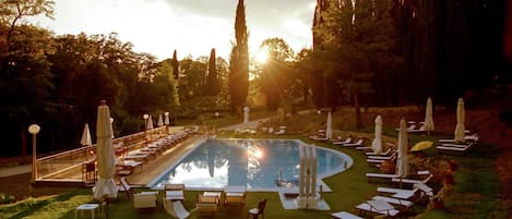 A pool area with lounge chairs and umbrellas, surrounded by trees and a clear sky.