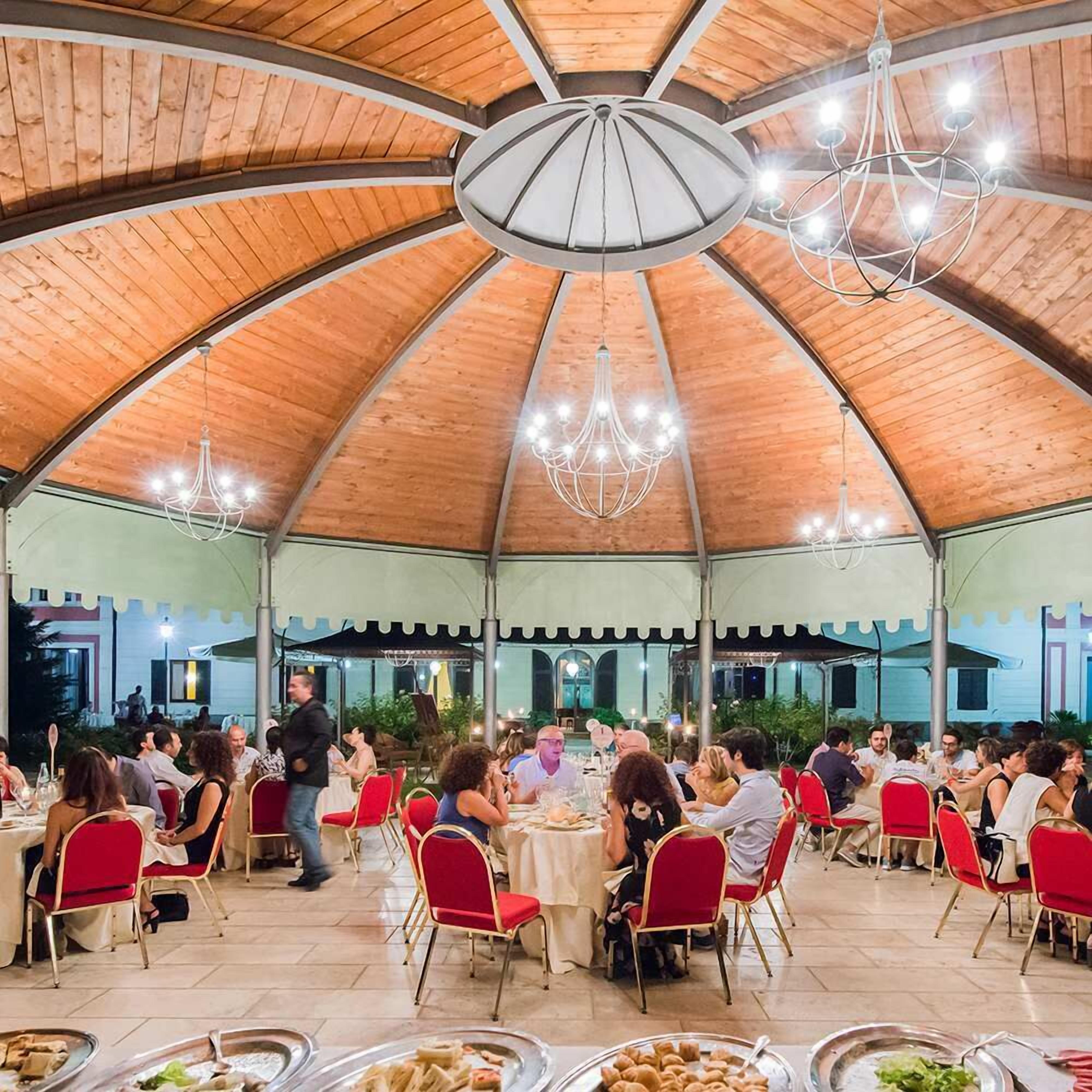 Uma área de refeições coberta com várias mesas e cadeiras, um lustre central e teto de madeira.