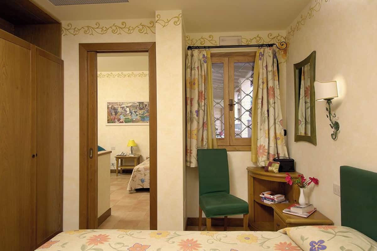 A bedroom with a bed, a chair, a small table with a lamp, a window with curtains, and a door leading to another room.