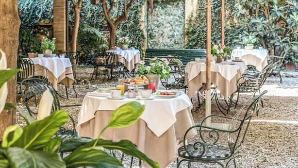 An outdoor dining area with round tables covered in white tablecloths, surrounded by metal chairs and shaded by umbrellas. - Hotel Santa Maria (Rome)