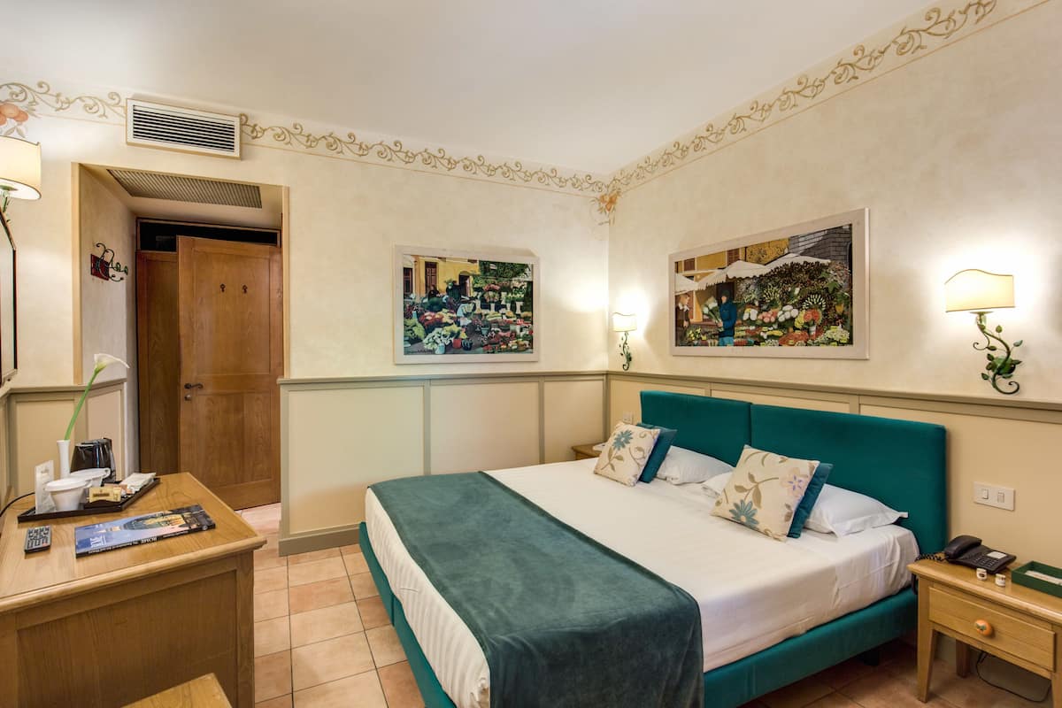A hotel room with a bed, bedside tables, a desk, and two wall-mounted paintings.