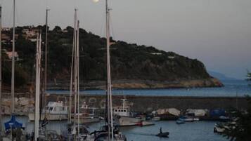 Una marina con diverse imbarcazioni ormeggiate, una costa rocciosa e una collina sullo sfondo con edifici.