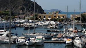 Un puerto deportivo con varias embarcaciones amarradas, un edificio con toldos amarillos y un paisaje montañoso al fondo.